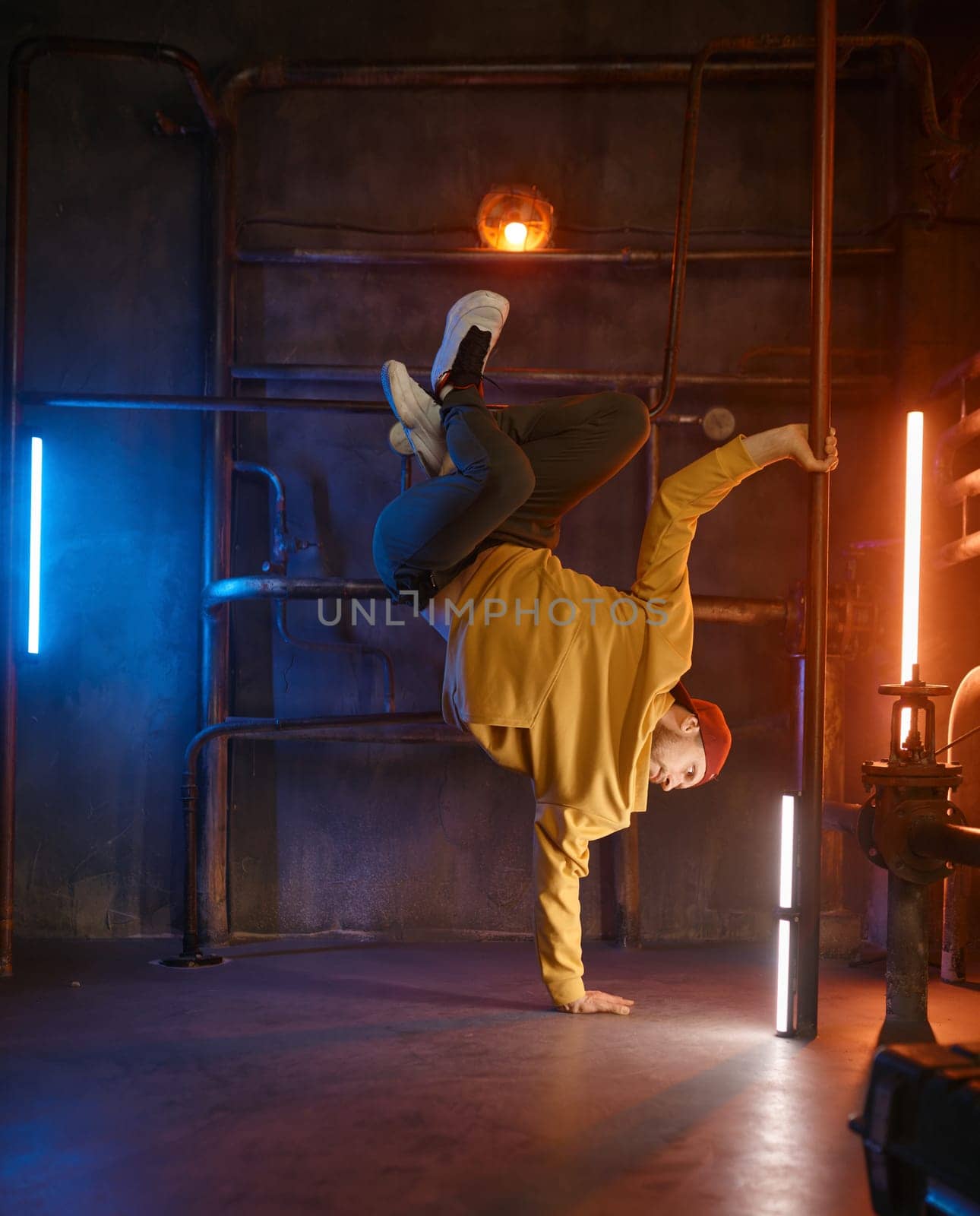 Modern hip hop dancer frozen in movie standing on one hand holding steel pilon tube practicing in dance over urban underground background