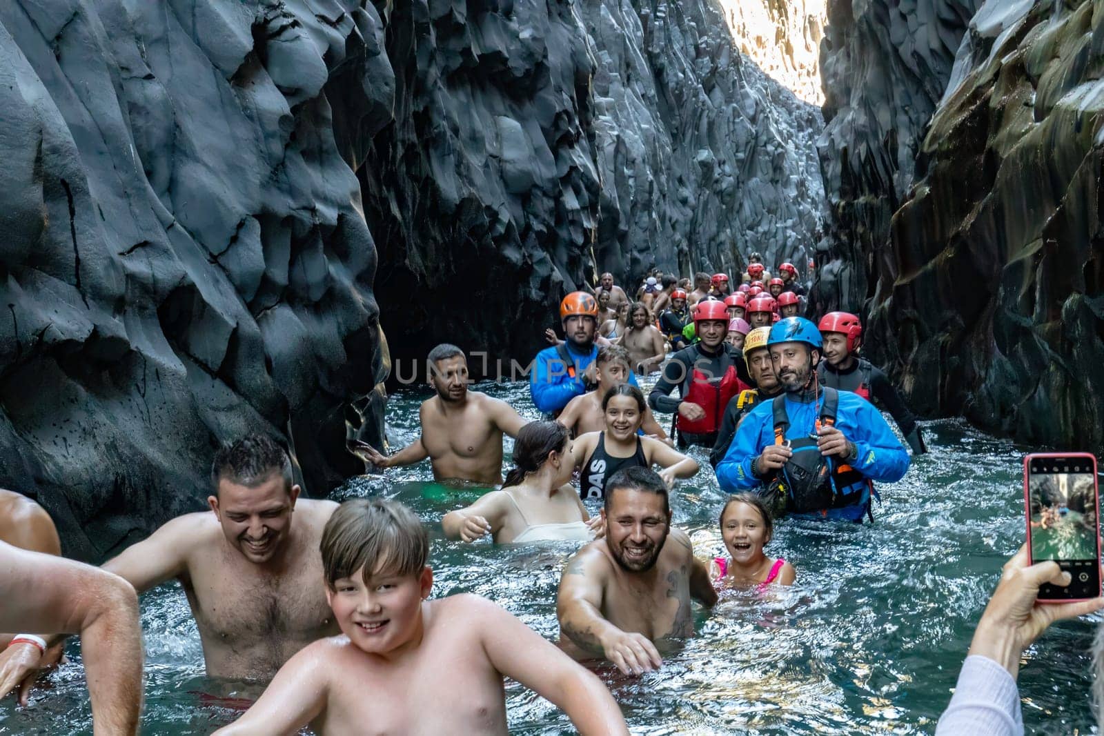 GOLE DI ALCANTARA, SICILY, ITALY - AUGUST 3 2022 diverse group of tourists passing through the cold river between the steep rocks of Gole Alcantara park