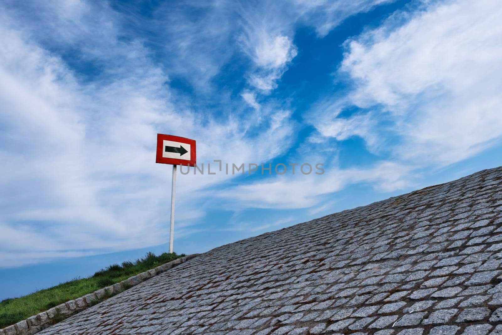 Black arrow on white background and in red frame standing atop of cobblestone paved hill at day. Blue sky with great clouds. Copy space, concept of direction, choice, signal, symbol, hope, options, opportunities, right