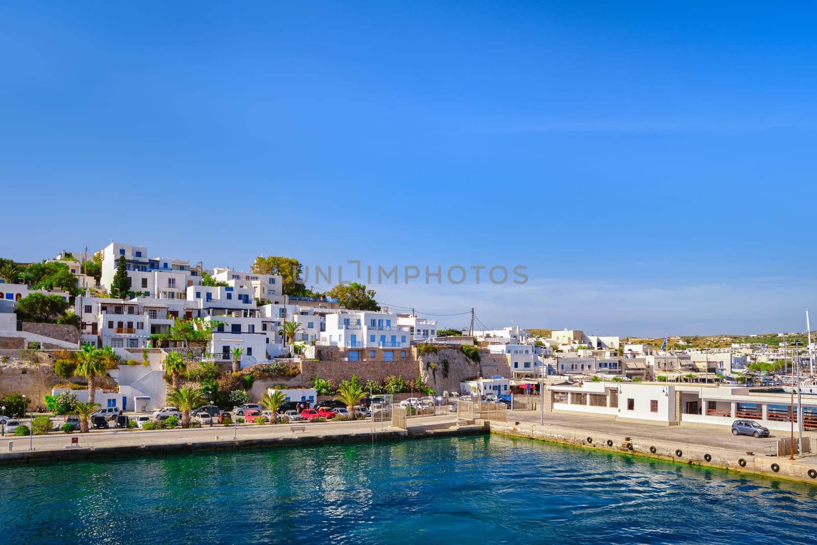 Beautiful summer day and sunshine in typical port town of Greek island. Whitewashed houses on hills and by waterfront. Mediterranean lifestyle, vacations, island hopping, crueses. Milos, Cyclades, Greece.