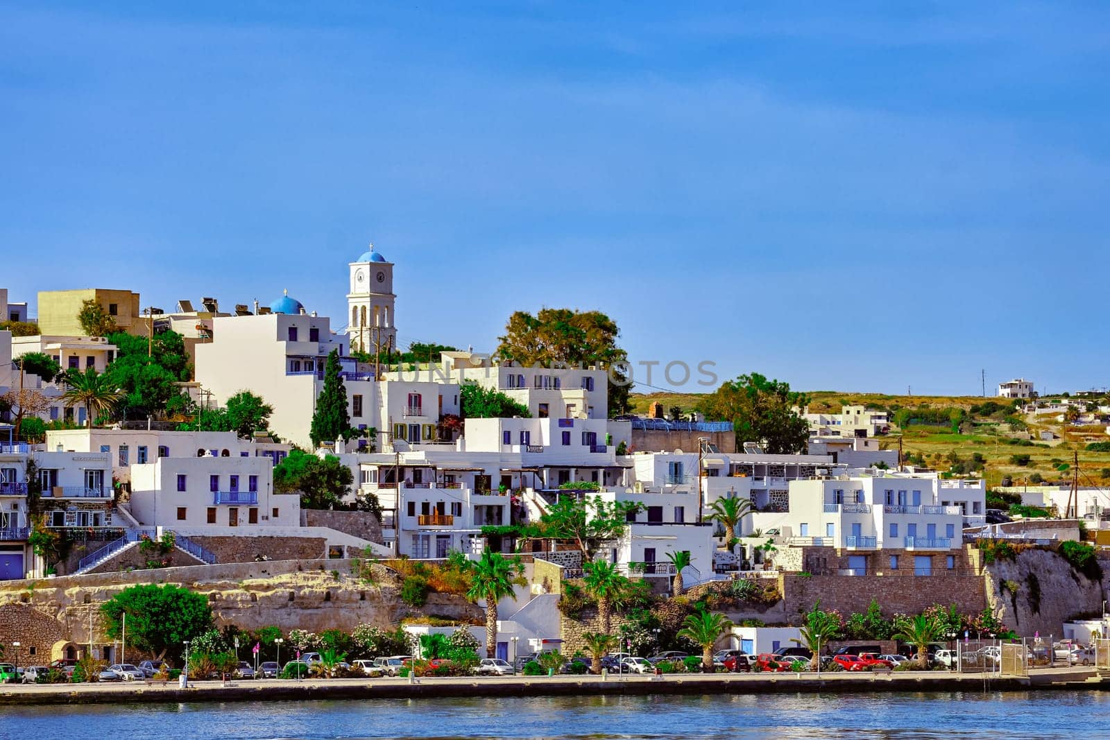 Beautiful summer day and sunshine in typical town of Greek island. Whitewashed houses on hills and by waterfront. Orthodox church. Mediterranean lifestyle, vacations. Milos, Cyclades, Greece.