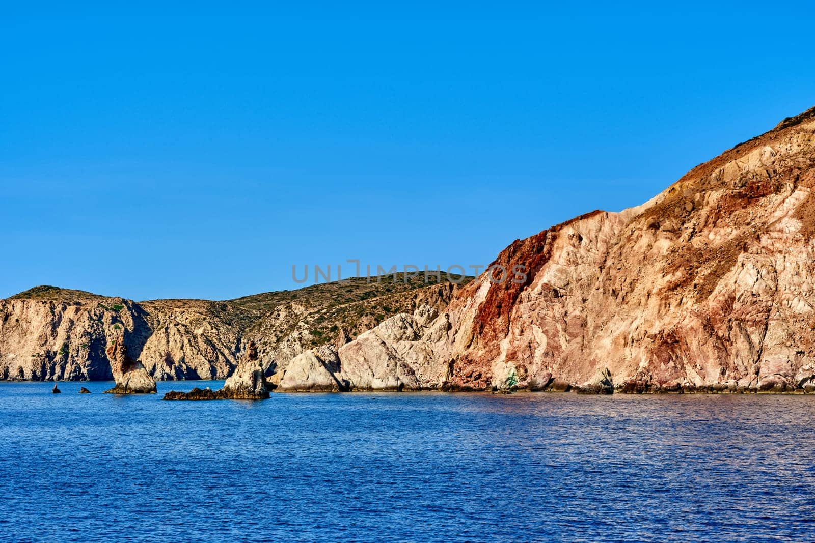 Beautiful view of high hill slope of island in Mediterranean Sea. Blue sea waters, clear blue sky, bright sunshine, summer day, rocks and islets of Aegean sea.