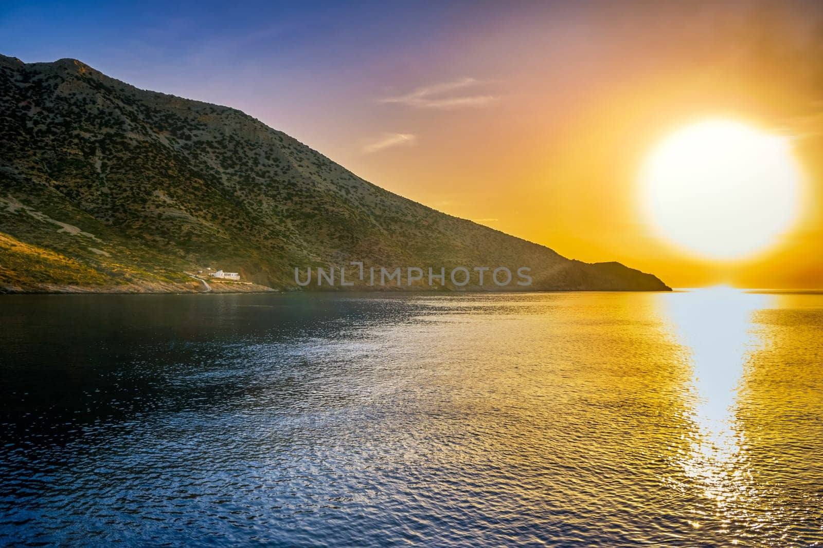 Beautiful view of Mediterranean island and its coast at sunset. Big low sun, sun flares and sun rays beam on lonely white Orthodox church on seashore. Milos island, Cyclades, Greece