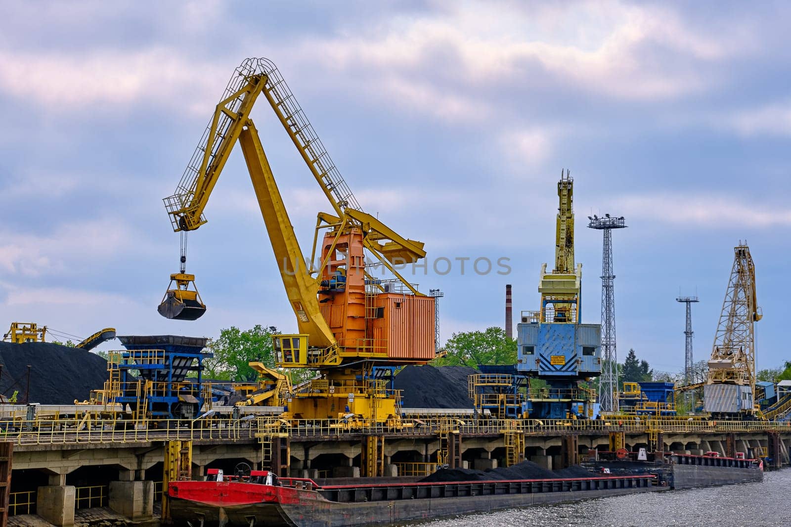 Close view of river port cranes with clamshell or griper loading coal to river drag boats or barges moored by pier on cloudy day. Problems of energy production, natural mineral deposits, environment crisis, alternatives