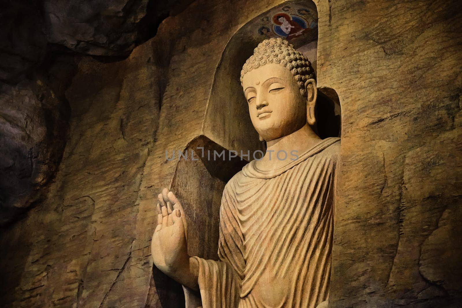 Statue of standing Buddha in monk clothes carved in limestone cave and lit by light. Right hand in adhay mudra gesture. Religion, tradition, prayer, meditation.