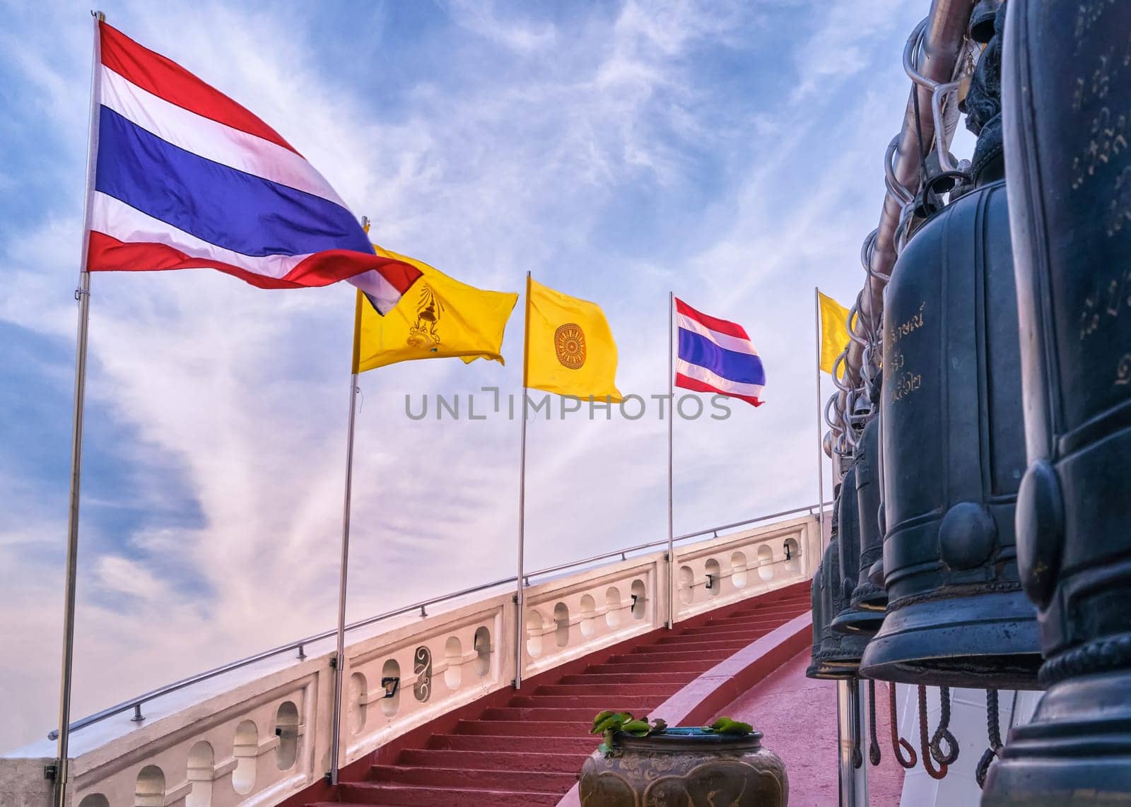 Row of Thai national, buddhism and Thai Royal flags along spiral staircase of red steps and white parapet, bells in blurred foreground. Stairway at Golden Mount temple or Wat Saket, Bangkok, Thailand. Incomplete inscriptions in Thai not translable.