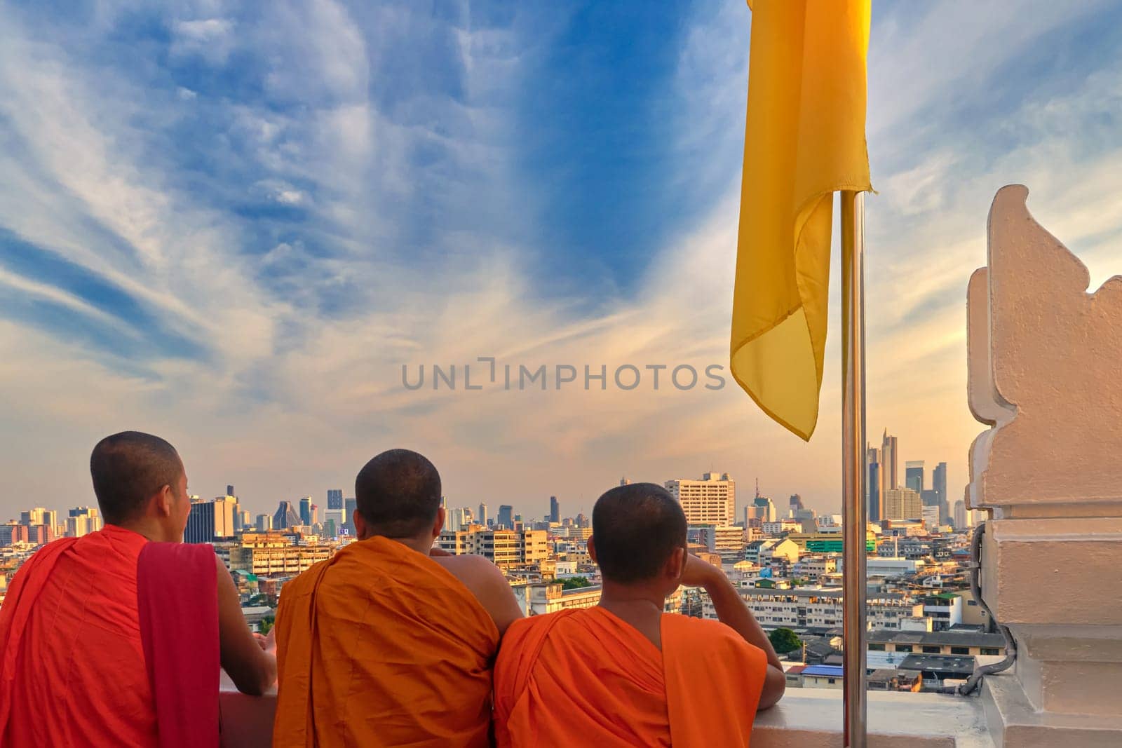 Three male Buddhist monks stand by white parapet wall of Wat Saket or Golden Mount temple and look at skyline of Bangkok, Thailand, at sunset. Orange robe, unrecognizable male adults, yellow flag, colorful cityscape, blue sky with clouds.