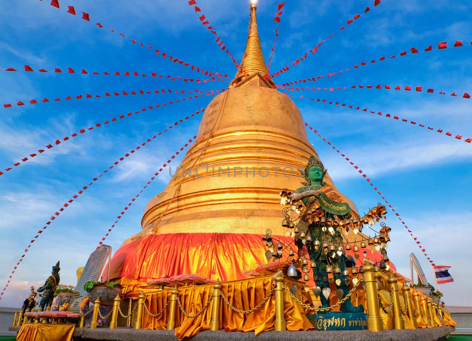 Statue of green-skinned Thai angel holding roll with message in Thai - translation: free from sorrow - stand in front of big golden stupa in sunlight and at blue sky with clouds in Buddhist temple. Decorations with bells, golden leaves and red flags.