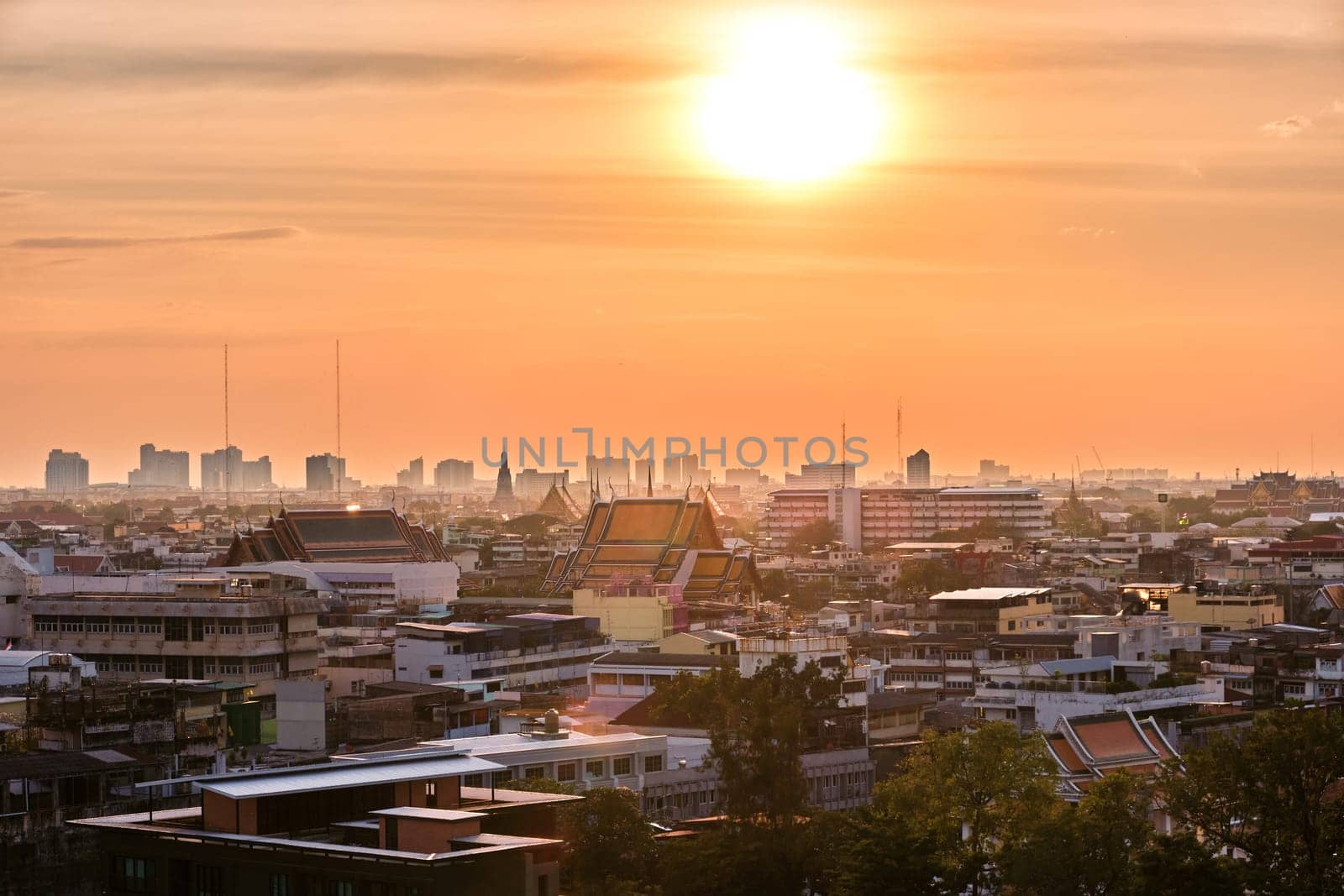 Beautiful cityscape of Bangkok city center in low sun, hazy orange sky and clouds, Thailand. Residential low-rise area in foreground, traditional temples, high-rises in downtown. Heat, summer, haze, heatwave, Asian climate, Buddhist religion.