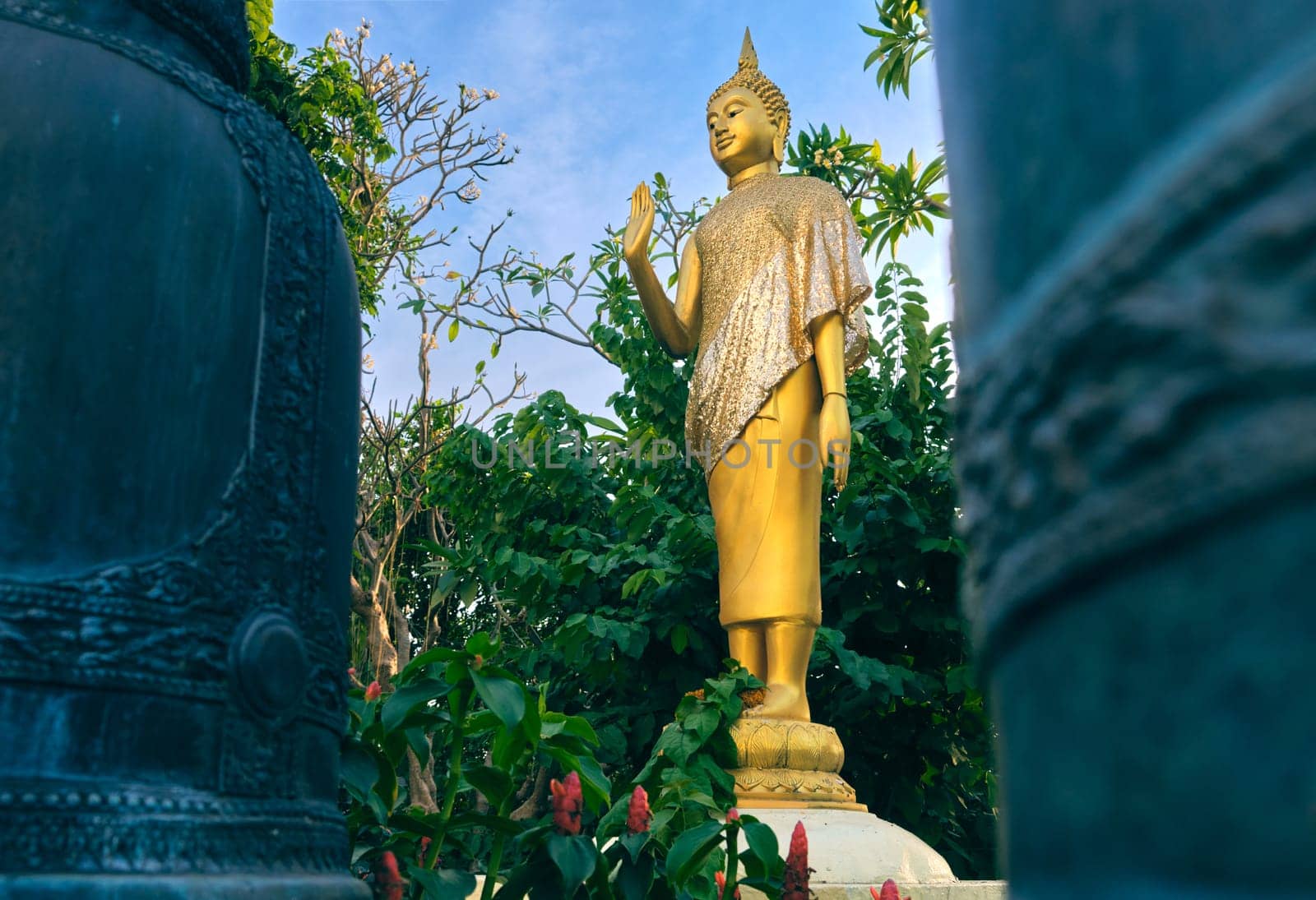 Gilded statue of standing Buddha in monk clothes and covered by golden textile shot between two bells and among trees and flowers. Right hand in adhaya mudra gesture. Selective focus, blurred foreground, religion, tradition, prayer, meditation.