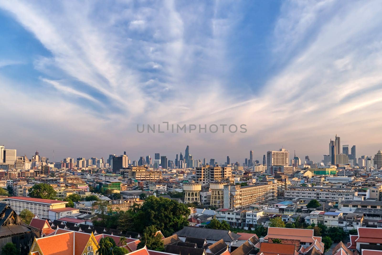 Beautiful cityscape of Bangkok city center in evening, low sun, blue sky and clouds, Thailand. Residential low-rises and traditional Buddhist temples in foregrounds, high-rises in downtown with King Power Mahanakhon skyscraper in distance. Colorful urbanscape