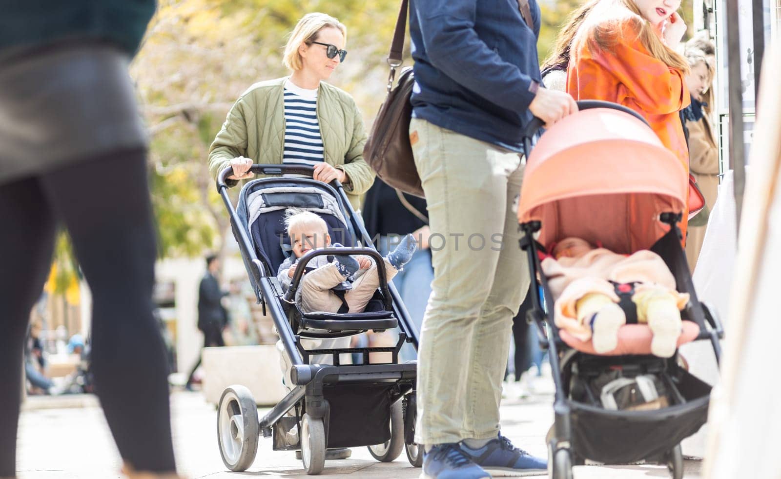 Mother walking and pushing his infant baby boy child in stroller in crowd of people wisiting sunday flea market in Malaga, Spain