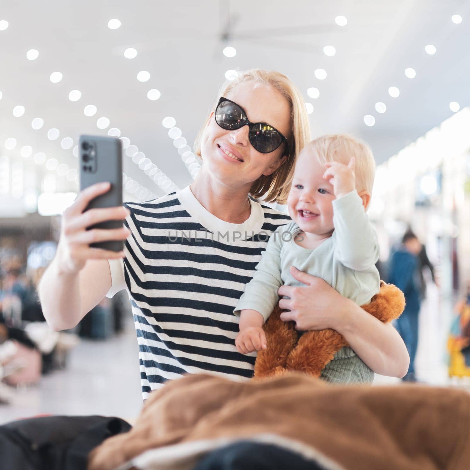 Mother taking selfie with mobile phone, while traveling with child, holding his infant baby boy at airport, waiting to board a plane. Travel with kids concept
