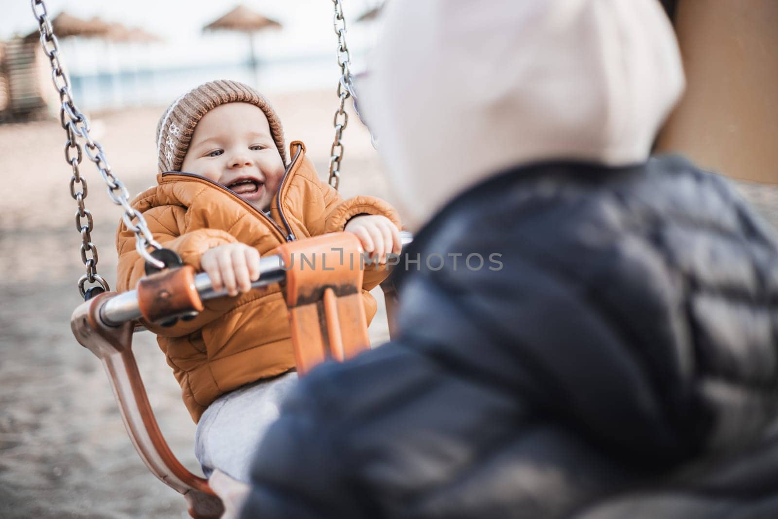 Mother pushing her cheerful infant baby boy child on a swing on sandy beach playground outdoors on nice sunny cold winter day in Malaga, Spain