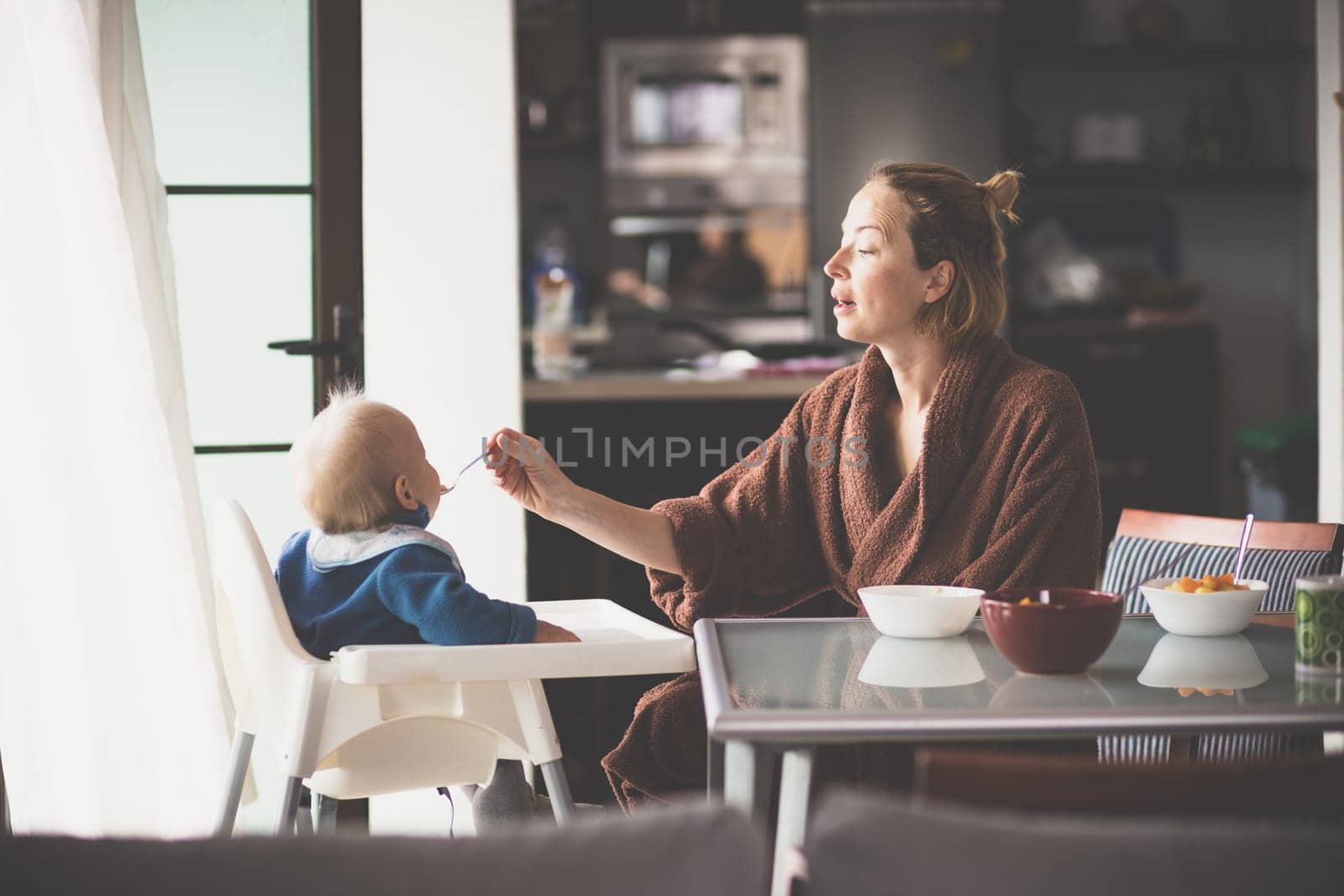 Mother wearing bathrope spoon feeding her infant baby boy child sitting in high chair at the dining table in kitchen at home in the morning.