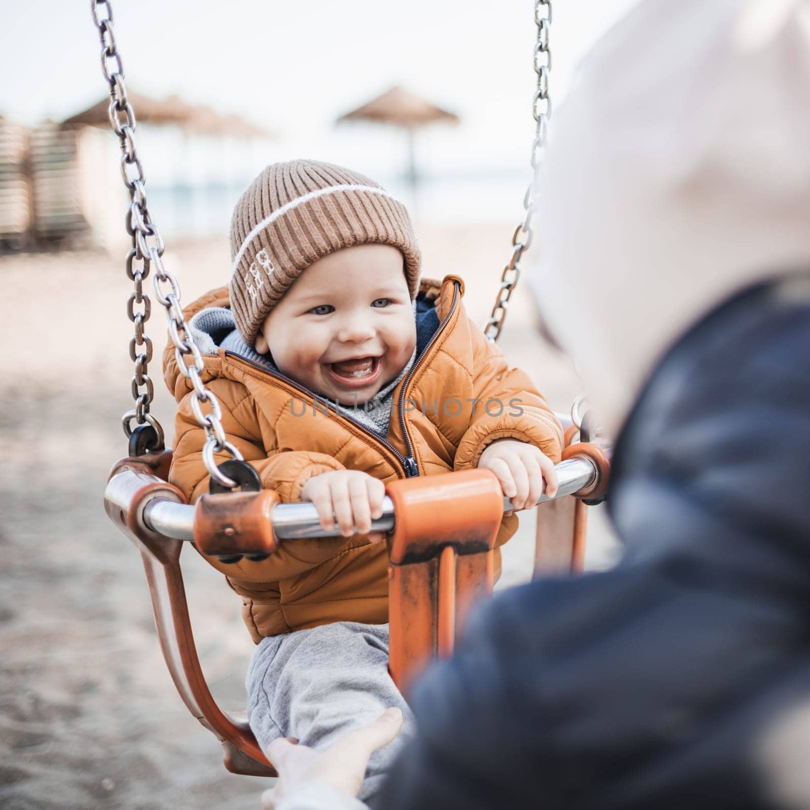 Mother pushing her cheerful infant baby boy child on a swing on sandy beach playground outdoors on nice sunny cold winter day in Malaga, Spain