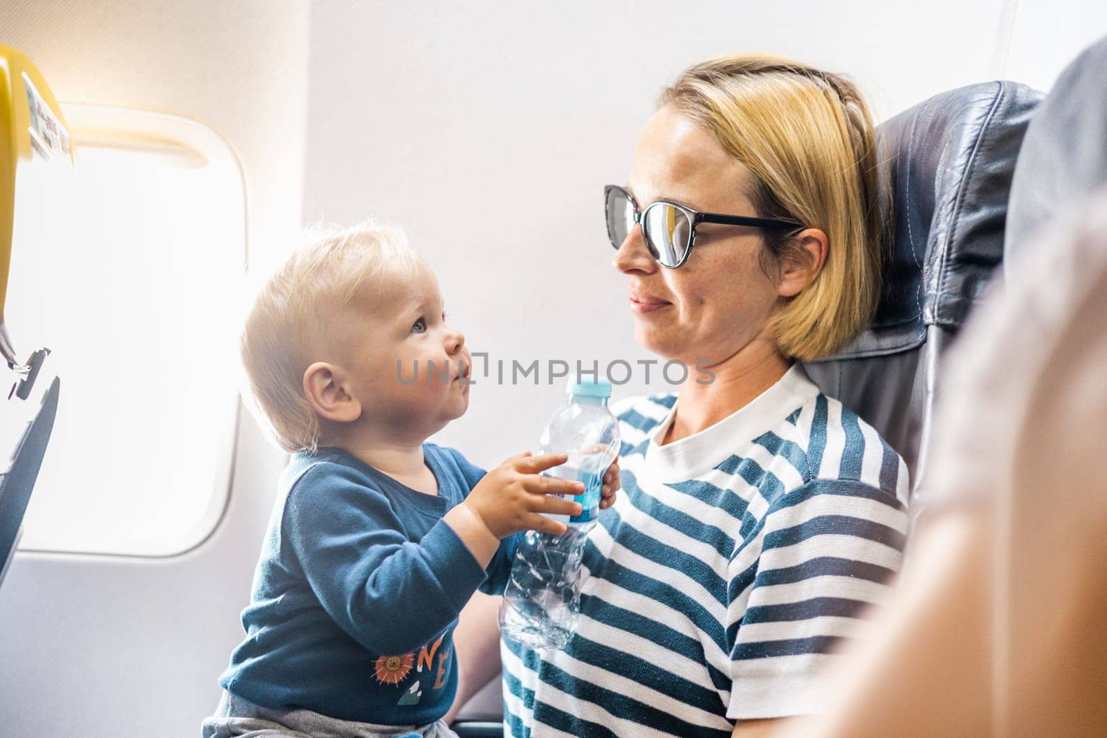 Mom and child flying by plane. Mother holding and playing with her infant baby boy child in her lap during economy comercial flight. Concept photo of air travel with baby. Real people