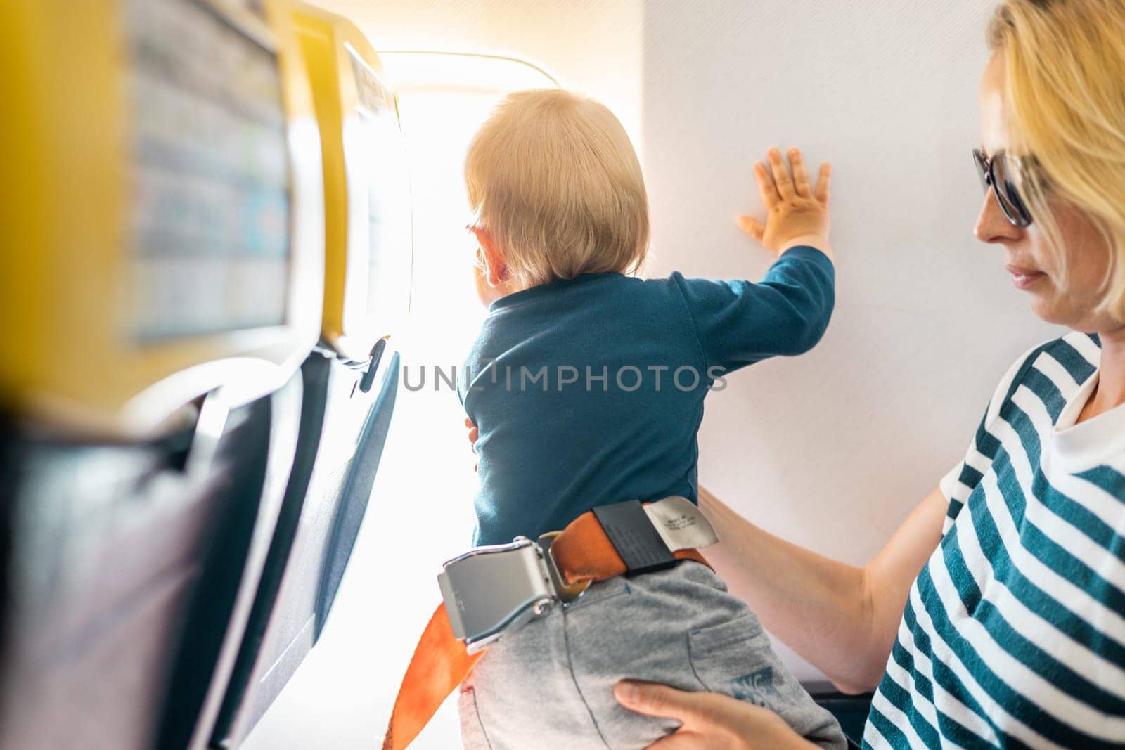 Mom and child flying by plane. Mother holding and playing with her infant baby boy child in her lap during economy comercial flight. Concept photo of air travel with baby. Real people