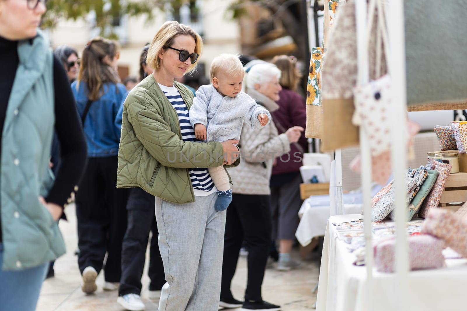 Mother walking carrying his infant baby boy child in crowd of people wisiting sunday flea market in Malaga, Spain.