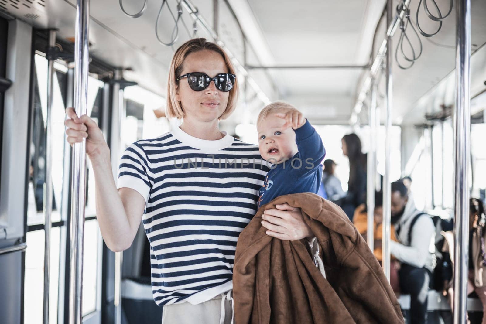 Mother carries her child while standing and holding on to bar holder on bus. Mom holding infant baby boy in her arms while riding in public transportation. Cute toddler traveling with mother