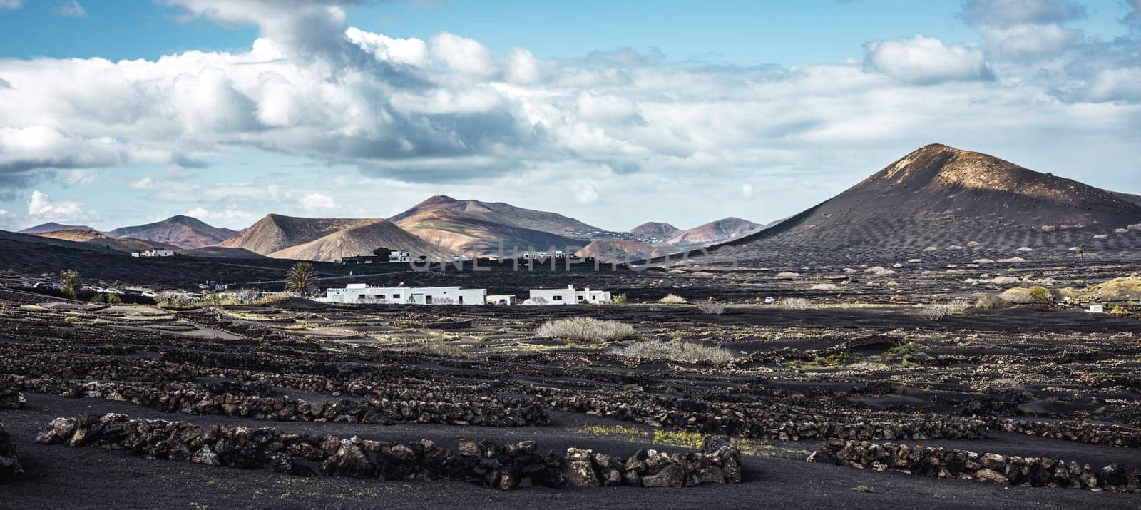Traditional white houses in black volcanic landscape of La Geria wine growing region with view of Timanfaya National Park in Lanzarote. Touristic attraction in Lanzarote island, Canary Islands, Spain