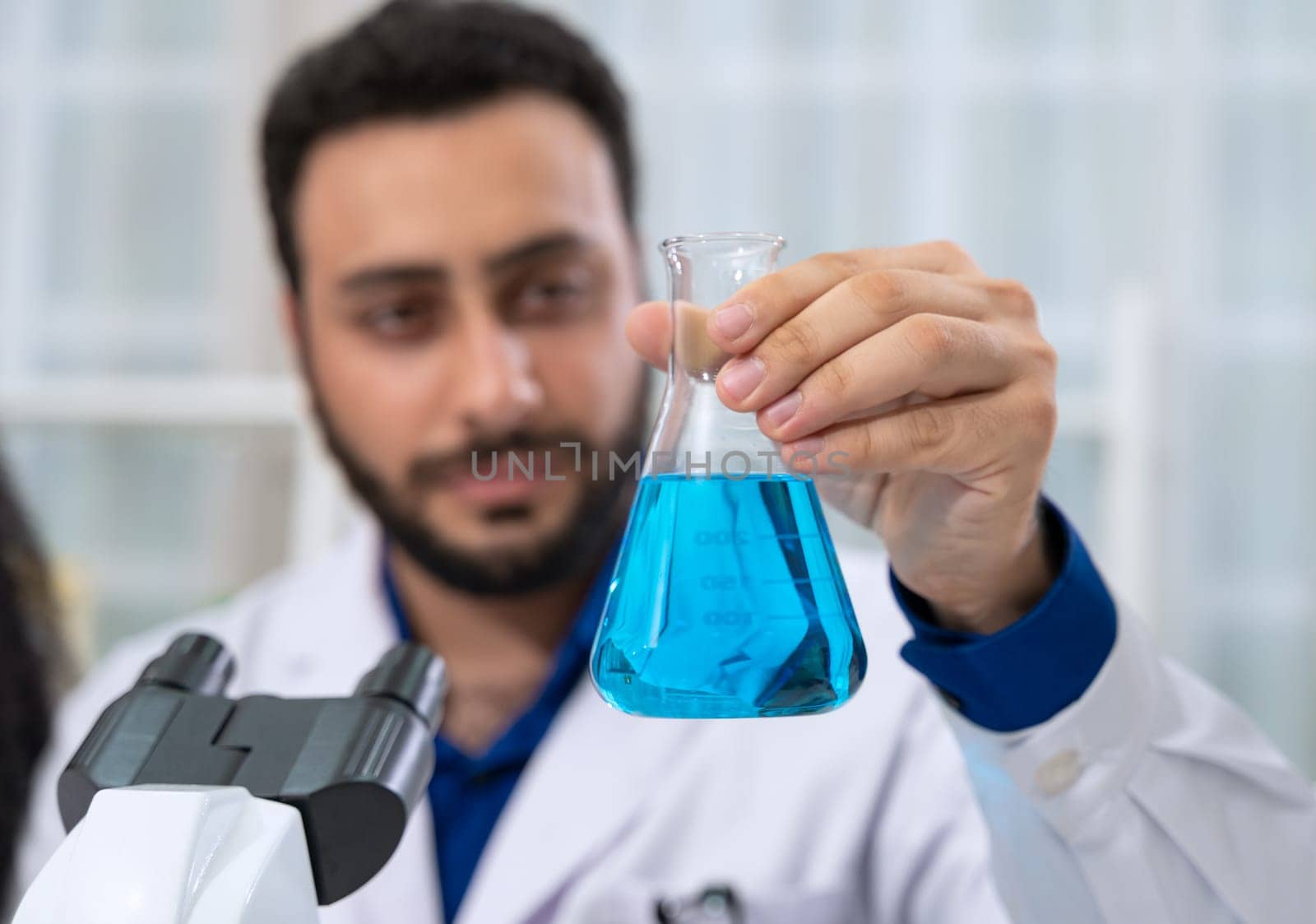 Scientist wearing white lab coat in research laboratory. Man with beard holding laboratory flask with blue reagent and using microscope