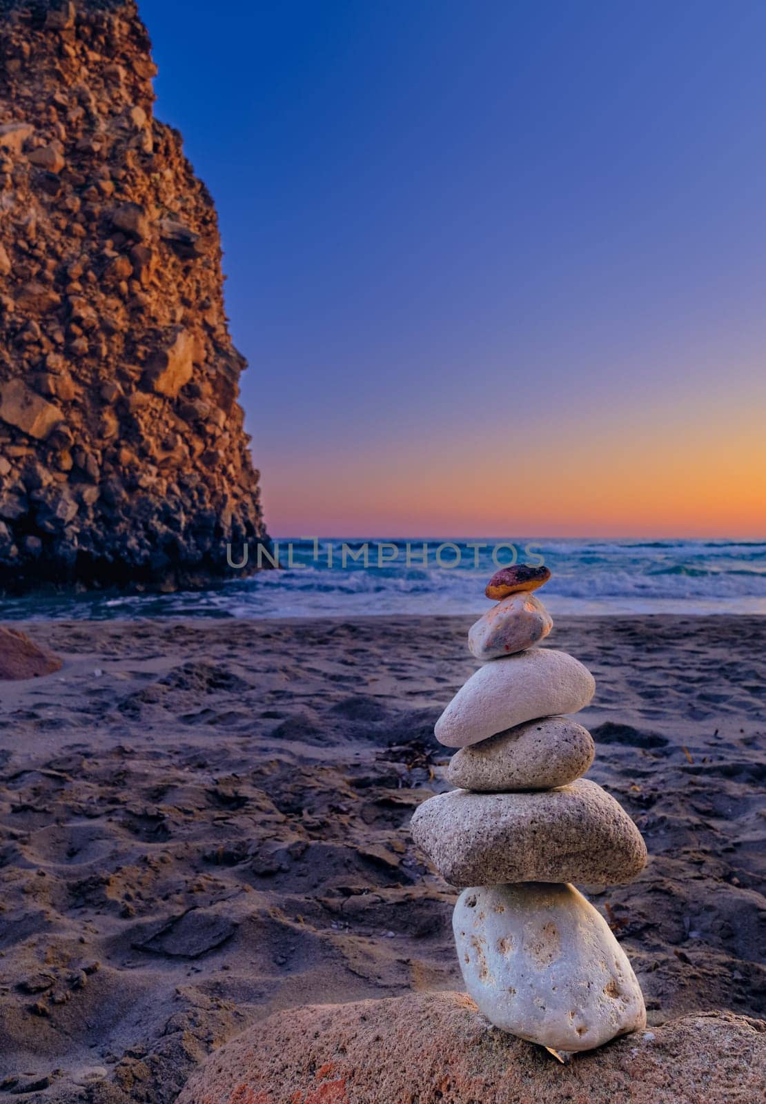 Small balance stone stack and big natural rock lit by sunset sun on sandy beach. Tidal waves in background, selective focus, blurred background, colorful view. Concept of balance, size, meditation, juxtaposition. Vertical shot