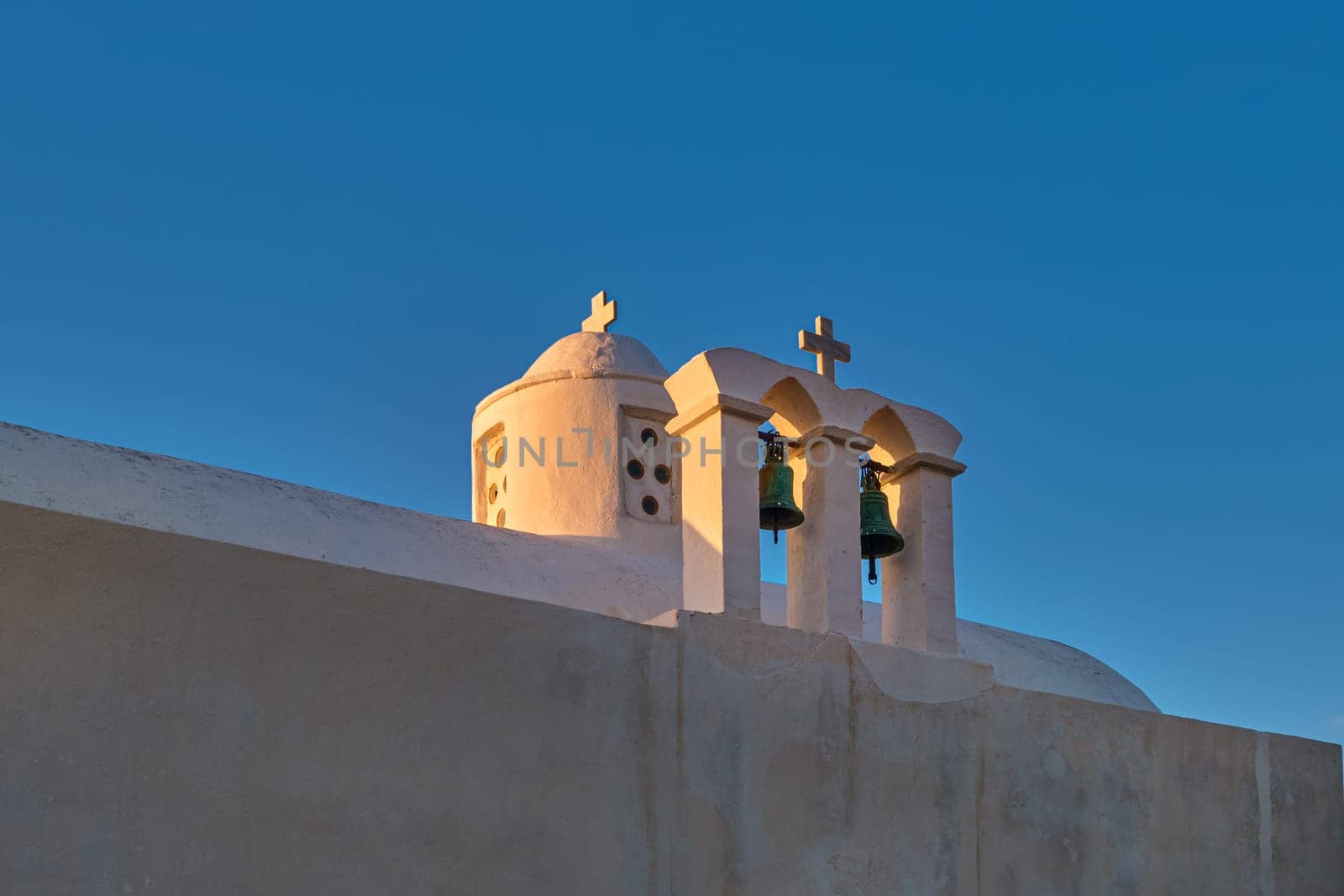 Whitewashed Greek Orthodox chapel, small belfry with cross and bell, roof and dome at sunset sky background. Christianity, religion, traditions, symbols, architecture. Close shot. Milos island, Greece