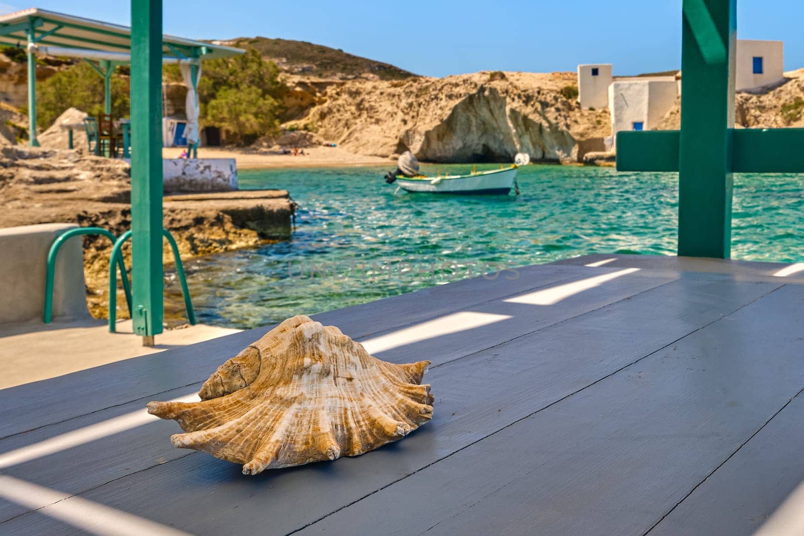 Summer vacations concept, conch sea shell on wooden table, open terrace by sea, Mediterranean island, bay, getaway landscape, Milos, Cyclades, Greece. Selective focus, blurred background.