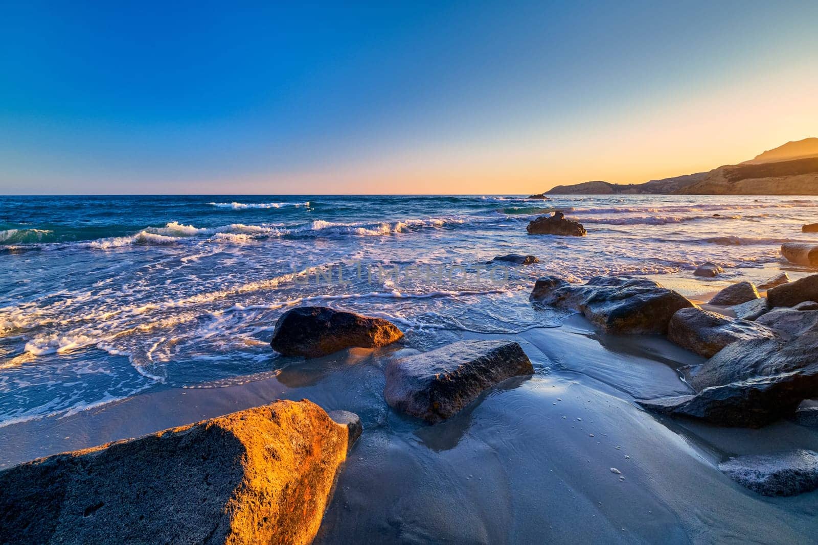Beautiful seascape of low tidal waves, clear sunset sky, hazy hills. Clear blue and azure waters approaching washed stone buried in sand. Nature, landscape, calm and relaxing meditative view. Mediterranean sea, Greece.