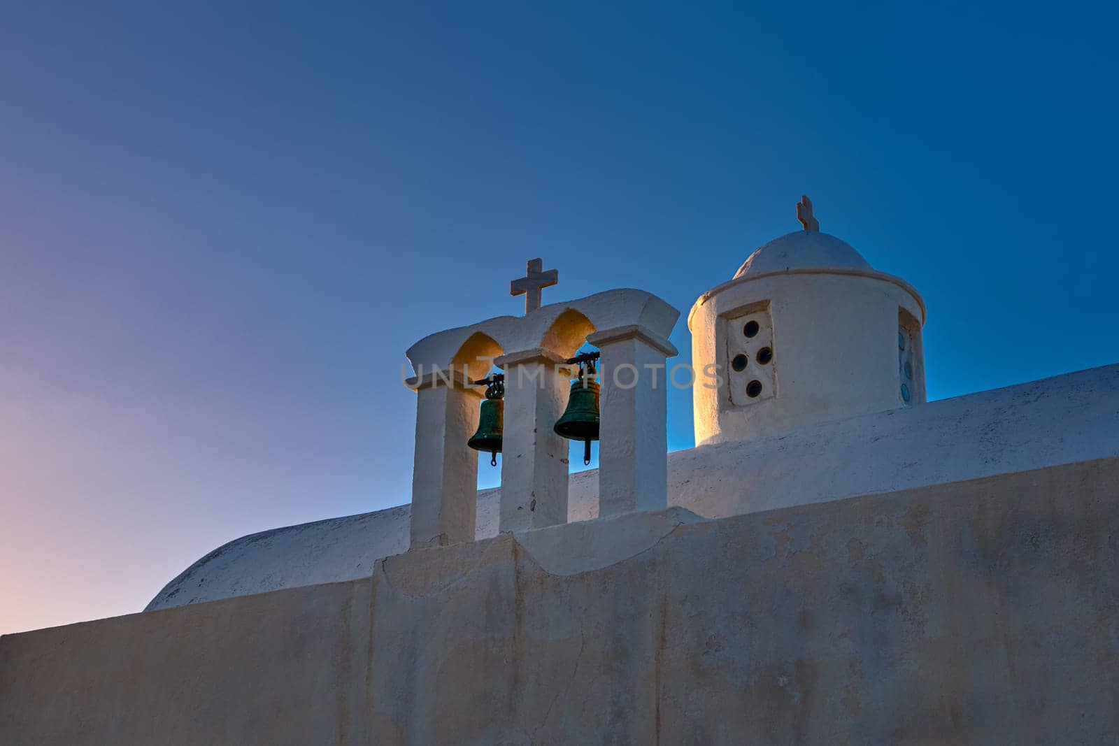 Whitewashed Greek Orthodox chapel, small belfry with cross and bell, roof and dome at sunset sky background. Christianity, religion, traditions, symbols, architecture. Close-up, diagonal shot. Milos island, Greece