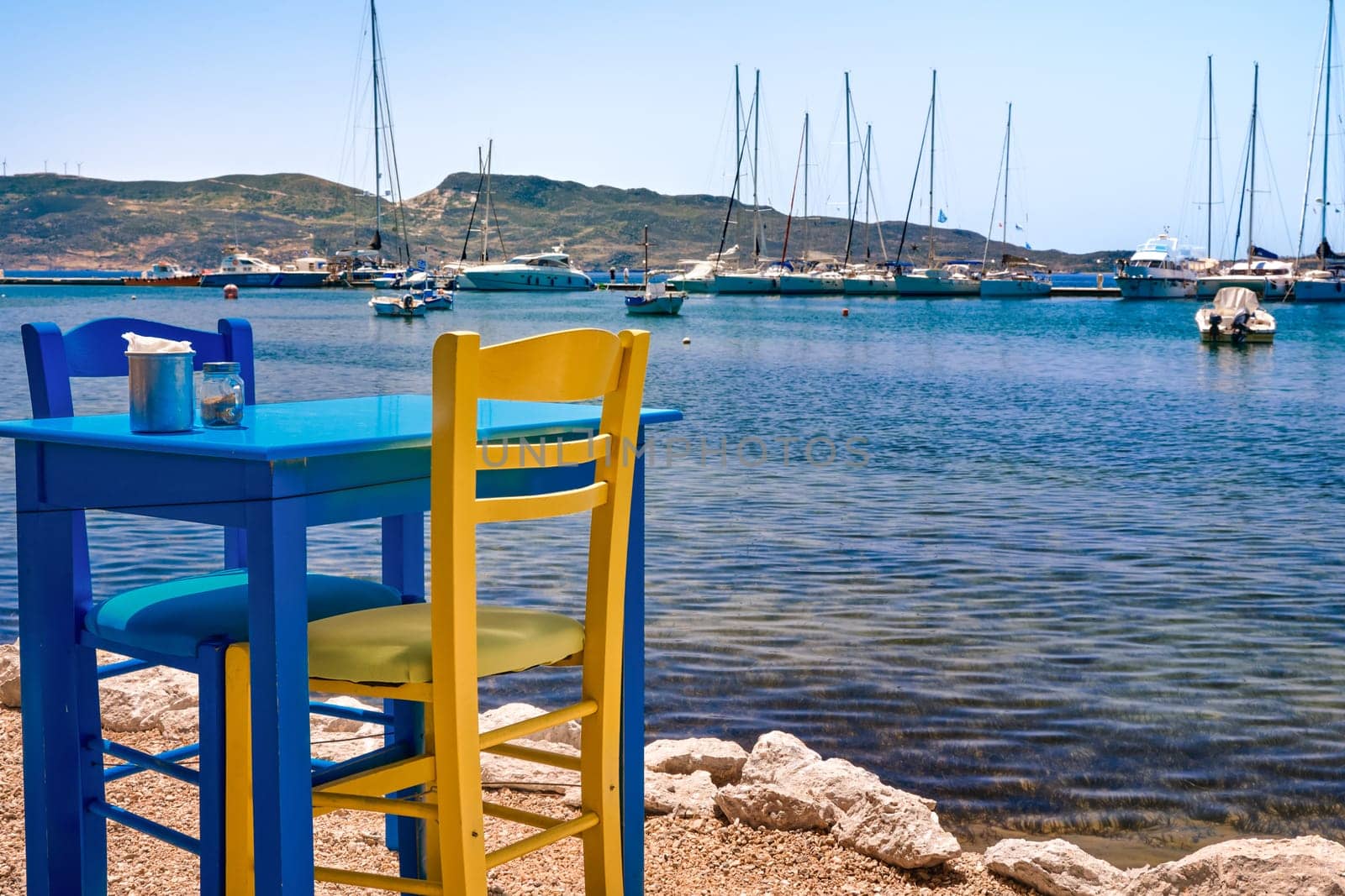 Colorful chair and table of Greek tavern by waterfront at sunny day. Vacations in Greece, summer, restaurant, outdoor dining, port, marina, yachts and boats, Mediterranean relax, Cyclades and Aegean islands. Selective focus, blurred background.