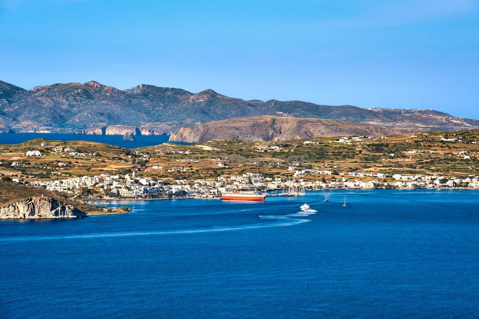 Beautiful summer day and sunshine in typical port town of Greek island. Whitewashed houses by waterfront, ferry moored by pier. Mediterranean lifestyle, vacations, island hopping, cruises. Milos, Cyclades, Greece.