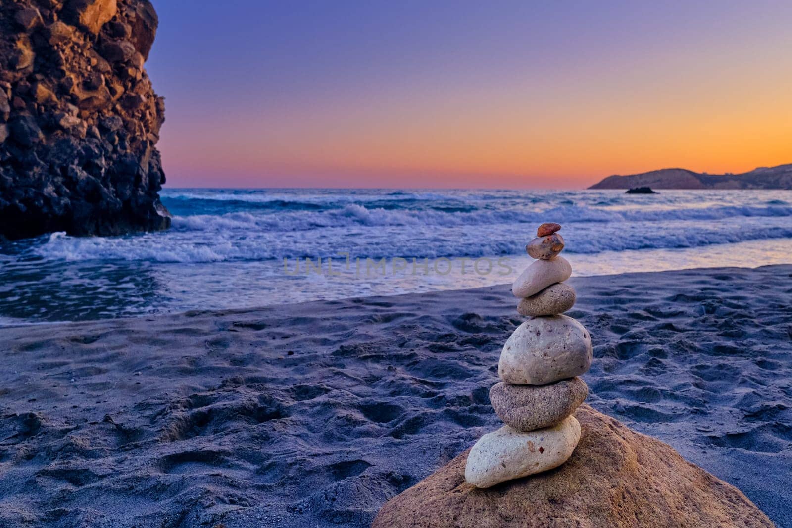 Small balance stone stack and big natural rock lit by sunset sun on sandy beach. Tidal waves in background, selective focus, blurred background, colorful view. Concept of balance, size, meditation, juxtaposition.