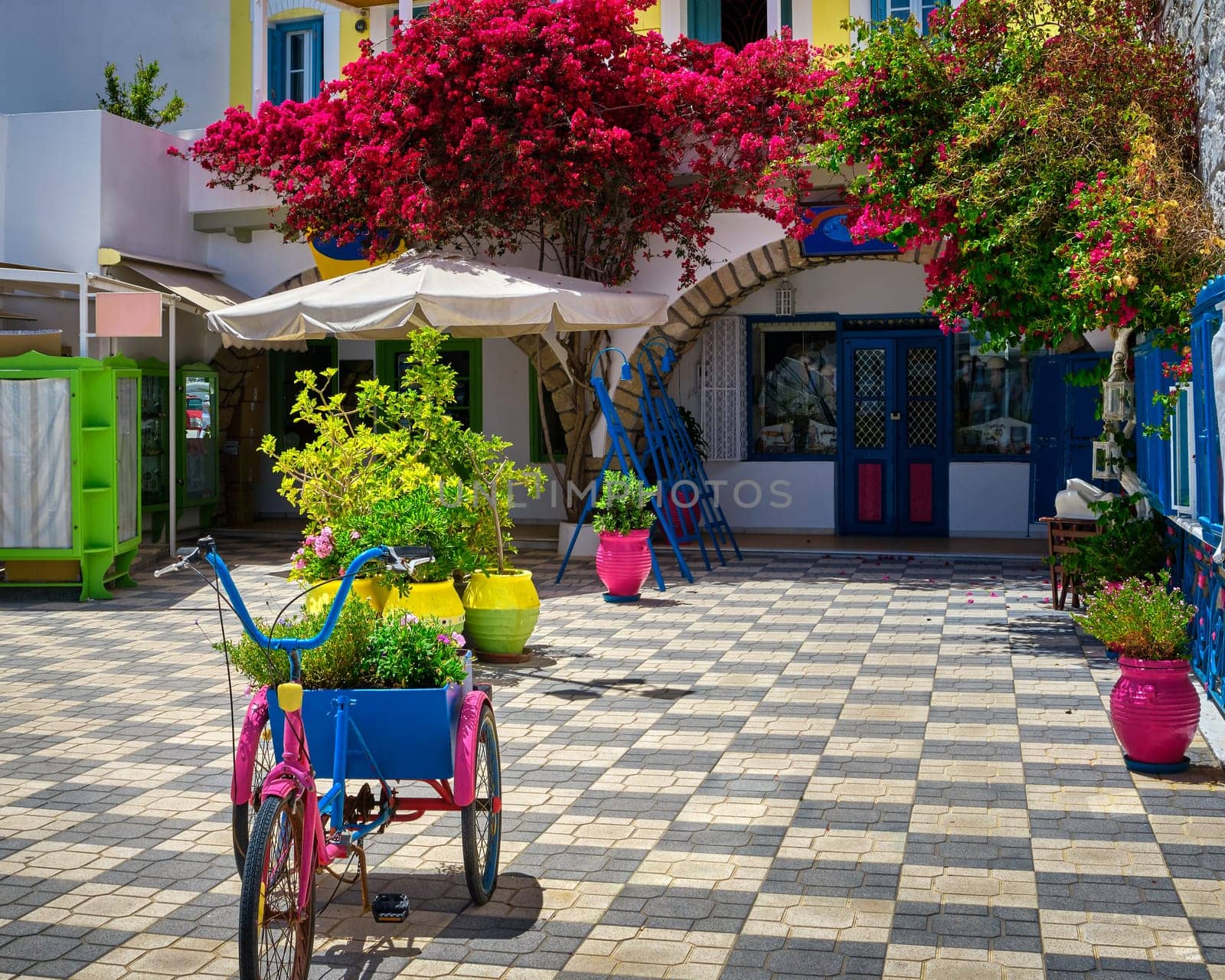 Beautiful corner shop in Adamas town, Milos, Cyclades islands. Greece. Summer sunshine, bougainvillea, flower pots, vines, small olive trees, cute colorful bicycle. Shop ready to open for tourists to enjoy the view, vacations, travels.