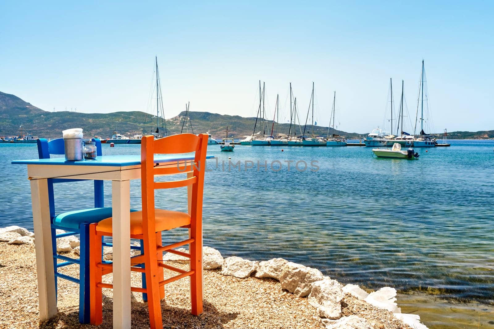 Colorful chairs and tables of Greek tavern by waterfront at sunny day. Vacations in Greece, summer, restaurant, outdoor dining, port, marina, yachts and boats, Mediterranean relax, Cyclades and Aegean islands. Selective focus, blurred background.