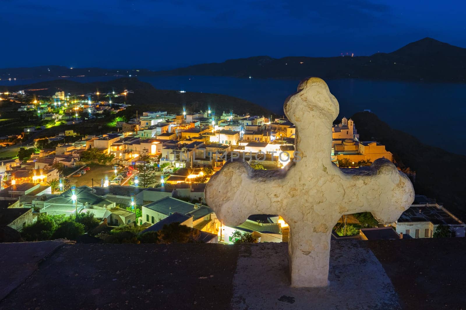 White Greek cross over colorful night view of traditional Greek small town or village by seafront, Plaka, Milos island, Cyclades, Greece. Whitewashed houses, Orthodox church, tiny lit-up streets, distant hills, bay waters, , Mediterranean lifestyle