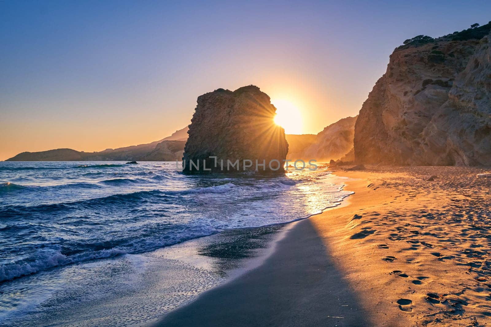 Colorful sunset over Fyriplaka beach, Milos island, Cyclades, Greece. Beautiful sky, unique lonely rock on sandy beach, low sun, sundown haze, rocky hills, sun rays, sea waves spray.
