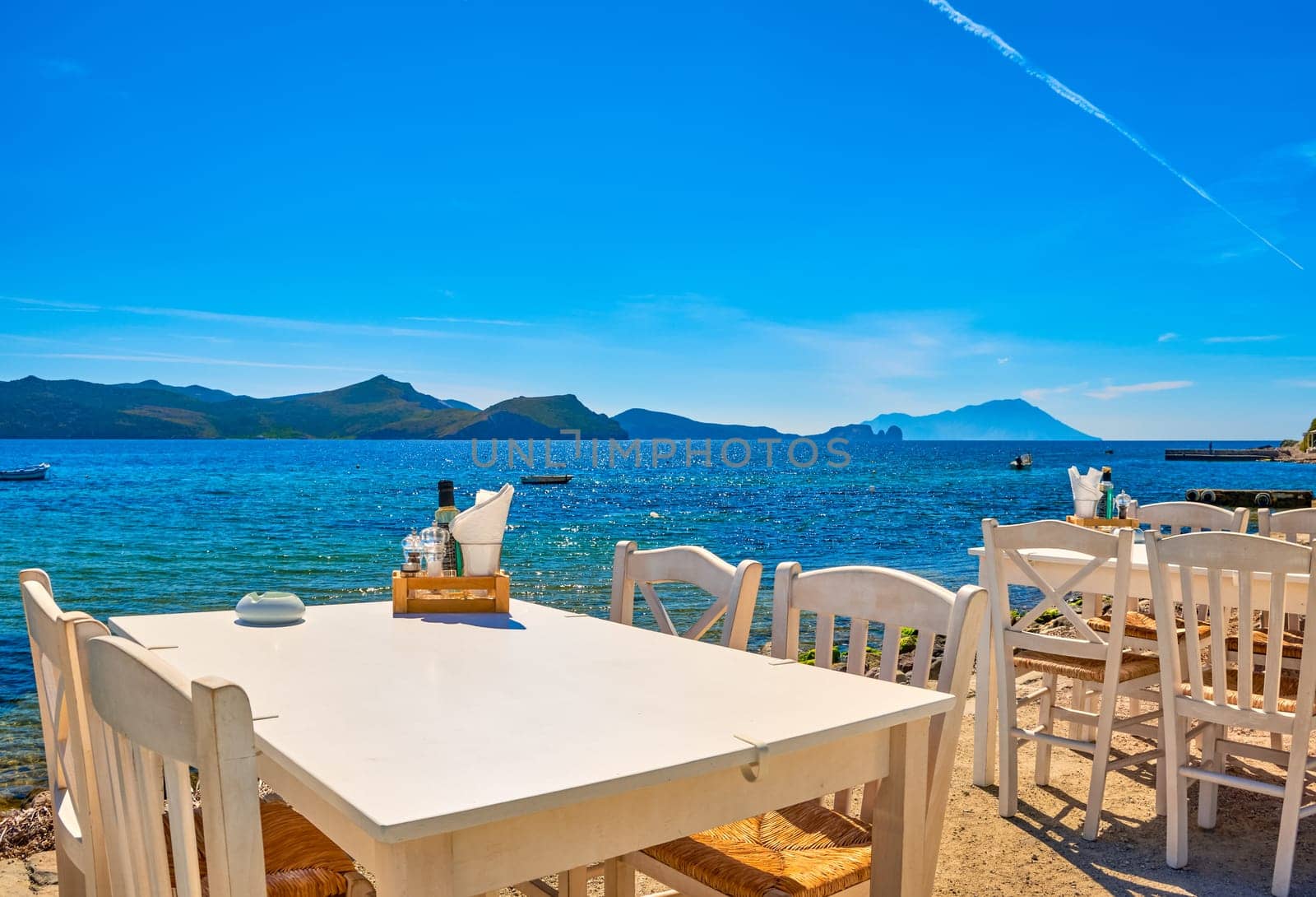 White chairs and tables of Greek tavern by waterfront at sunny day. Vacations in Greece, summer, restaurant, outdoor dining, port, marina, yachts and boats, Mediterranean relax, Cyclades and Aegean islands. Selective focus, blurred background.