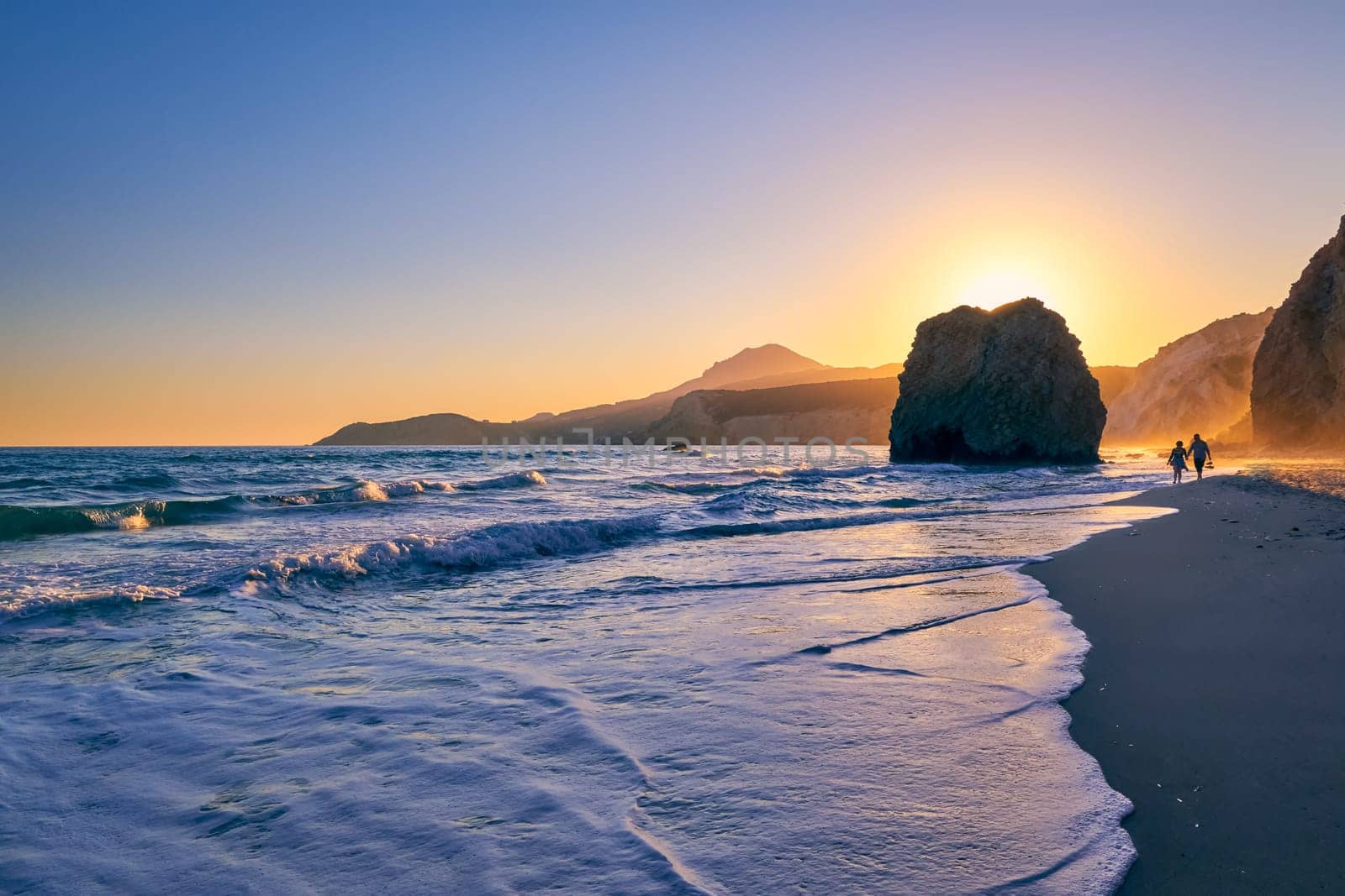 Distant silhouettes of two adults walking in tidal waters on Fyriplaka beach, Milos island, Cyclades, Greece. Beautiful sunset sky, unique lonely rock on sandy beach, low sun, sundown haze, rocky hills.