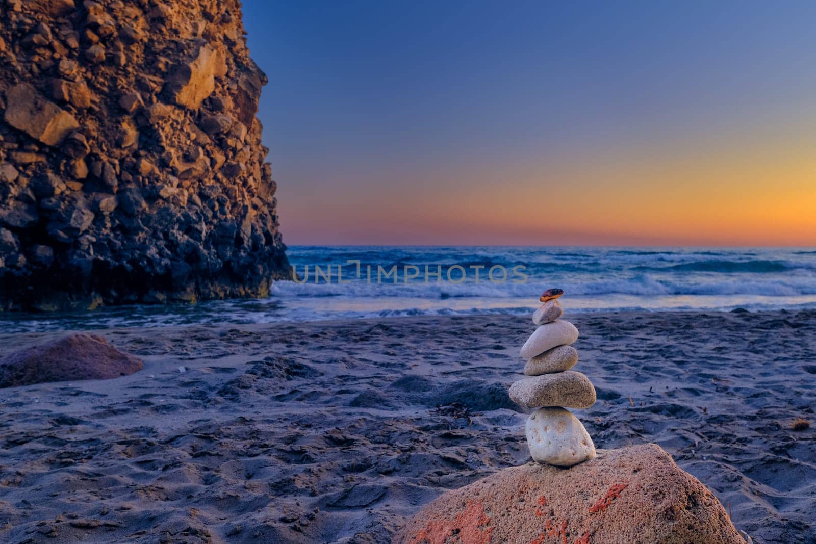 Small balance stone stack and big natural rock lit by sunset sun on sandy beach. Tidal waves in background, selective focus, blurred background, colorful view. Concept of balance, size, meditation, juxtaposition.