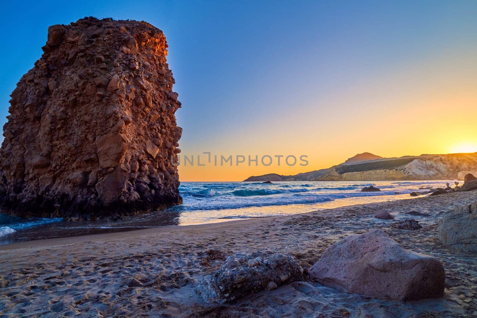 Colorful sunset over Fyriplaka beach, Milos island, Cyclades, Greece. Beautiful sky, unique lonely rock on sandy beach, low sun, sundown haze, rocky hills, sun rays, sea waves spray.