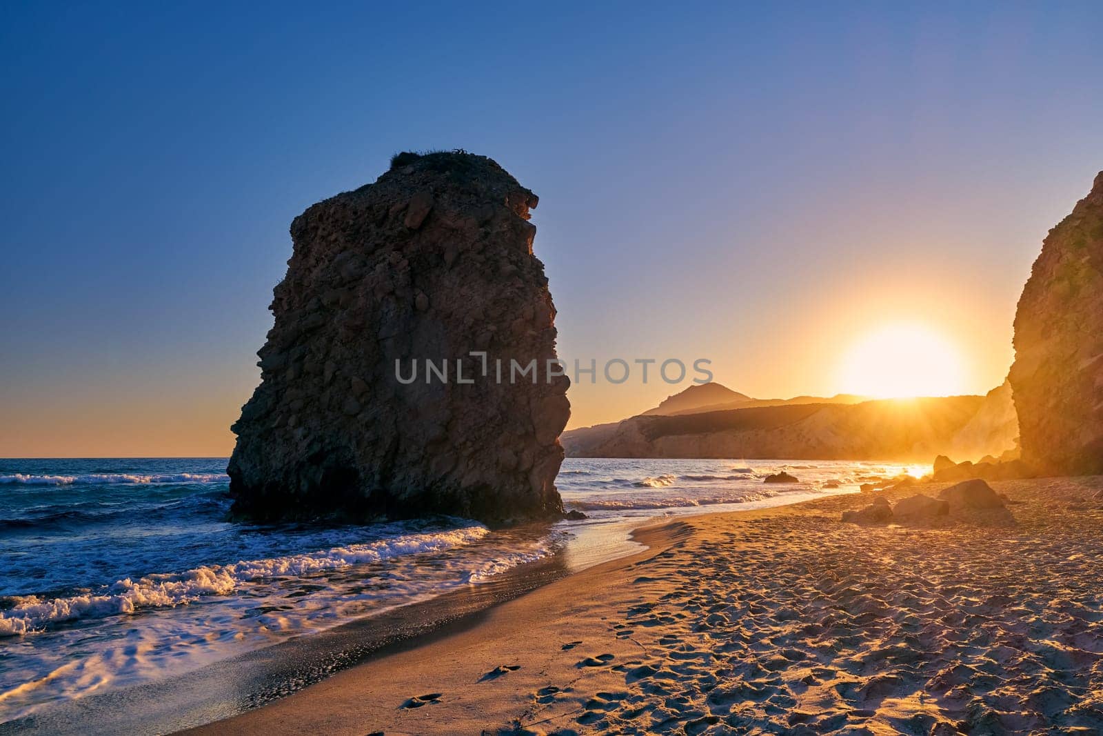 Colorful sunset over Fyriplaka beach, Milos island, Cyclades, Greece. Beautiful sky, unique lonely rock on sandy beach, low sun, sundown haze, rocky hills, sun rays, sea waves spray.