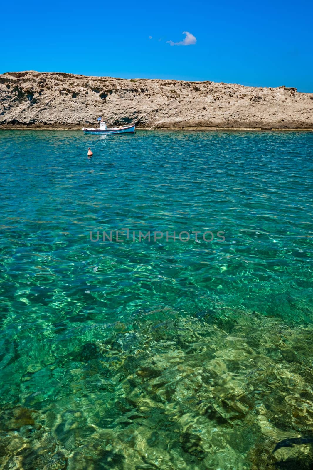 Anchored small boat under Greek flag, azure sea waters, clear blue sky, rocky hill, red fishing buoy floating. Frontal shot. Milos, Greece. Summer vacations background, concept, copy space, no people, vertical composition, book cover.