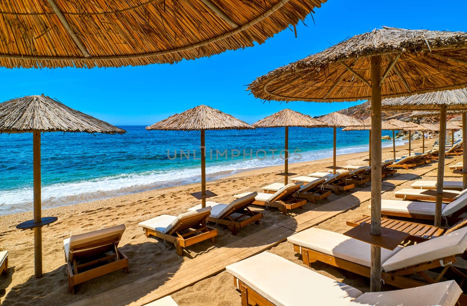Two rows of sun umbrellas and sunbeds ready for tourists on sandy beach at day sunshine. Perfect summer destination. Straw sunshades and sunbeds on the empty beach with blue sea and rocky hills. Vacations in Greece, islands retreat. Milos, Greece.