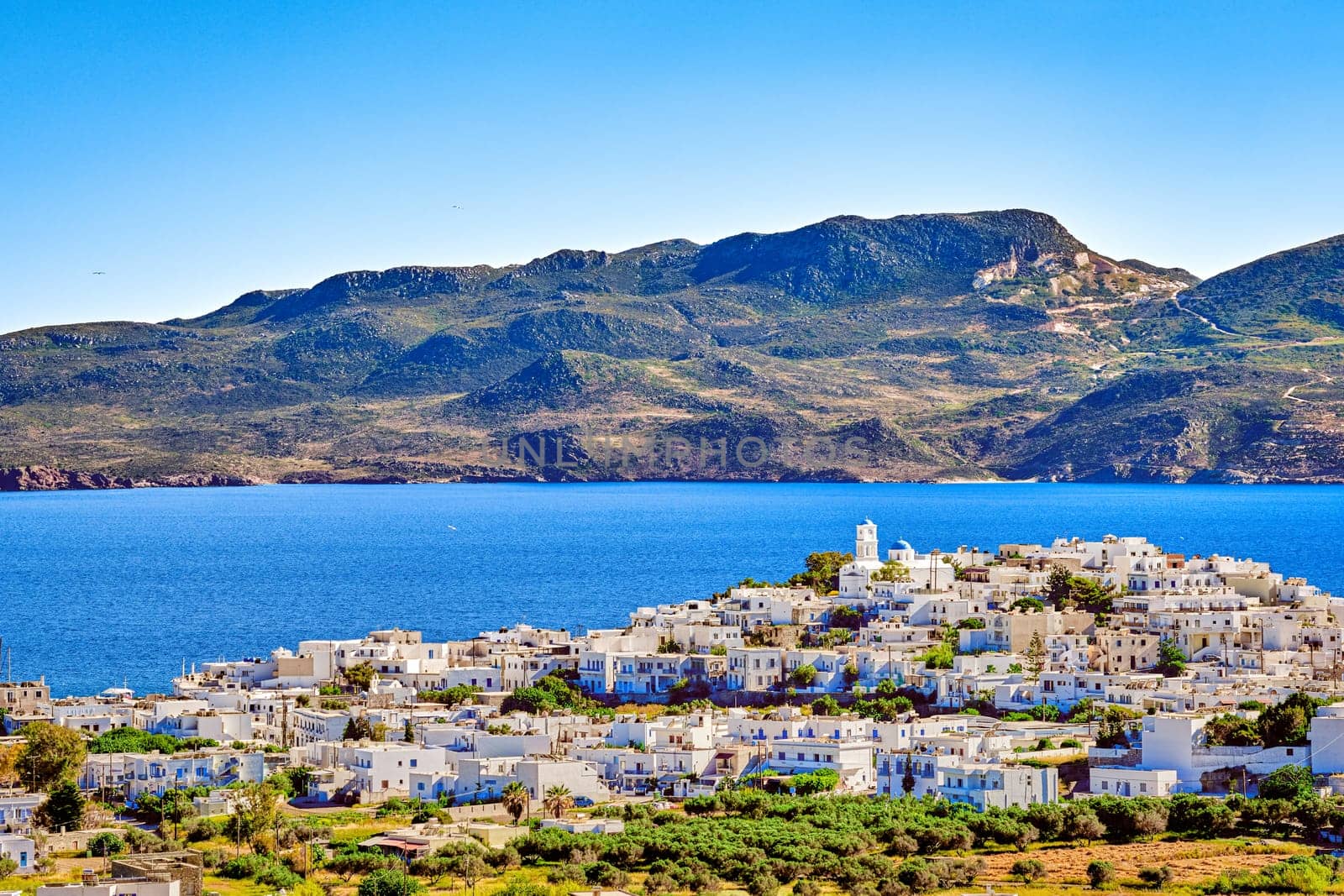 Beautiful view of Adamantas, main town of Milos island, Greece, on sunny day. Hill top town, whitewashed houses, church bell tower, blue sea waters of bay and distant hazy hills. Greek islands, Mediterranean sea, summer sunshine.