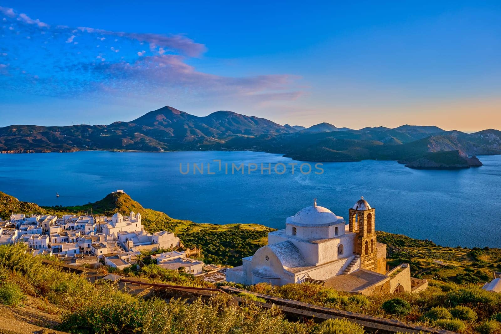 Beautiful view of Mediterranean island at sunset. Whitewashed Greek Orthodox church by seafront in rays of low sun. Sun glare, distant hills, blue sea, clear sky, distant hills, whitewashed houses, summer, vacations, Milos, Cyclades, Greece.