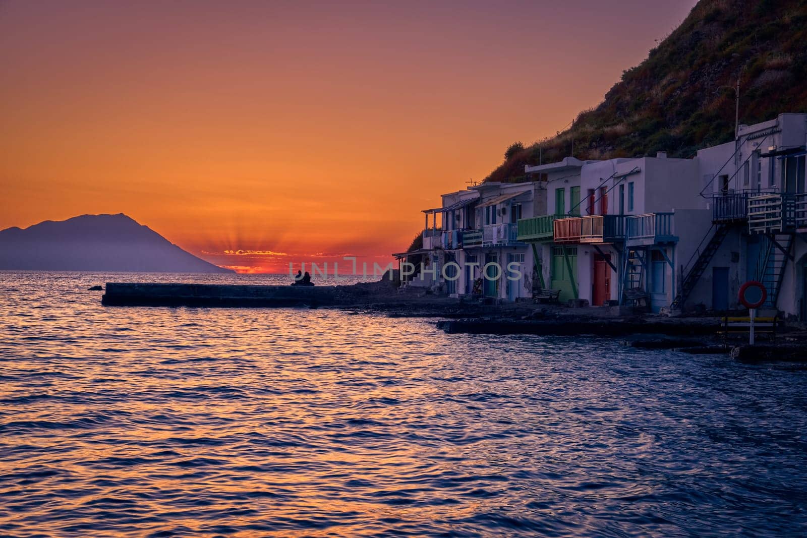 Beautiful view of sunset in Klima village, Milos island, Greece. Pair of adults enjoy sunset view on pier of small fishermen's village. Colorful houses by waterfront, distant mountains, calm sea, clear sky, romantic weekend, relax, vacations, voyage