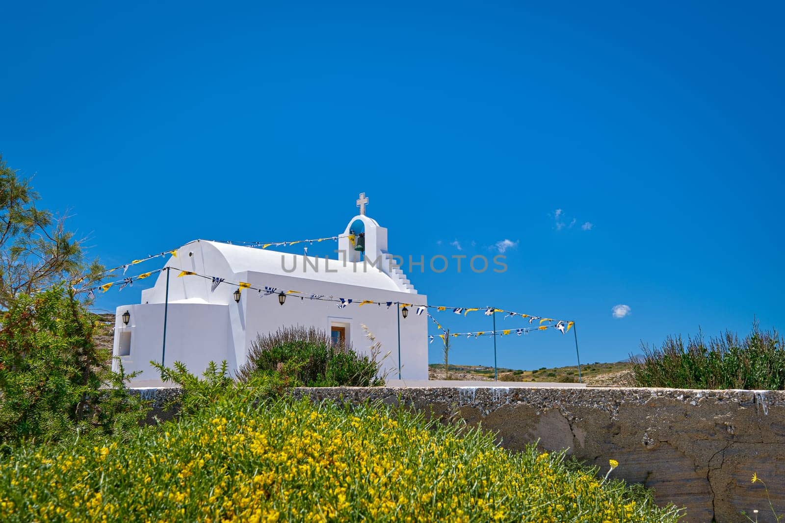 White Greek Orthodox chapel or church on hilltop of seashore against clear blue sky on sunny day. Traditional religious building. Colorful flowers and bushes in foreground. Typical Greek landscape. Diagonal composition, copyspace. Milos Greece