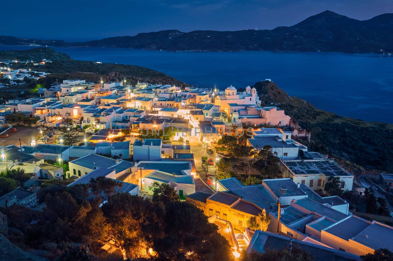 Colorful night view of traditional Greek small town or village by seafront, Plaka, Milos island, Cyclades, Greece. Whitewashed houses, Orthodox church, tiny lit-up streets, green hills, calm bay waters, hazy distant mountains, Mediterranean lifestyle